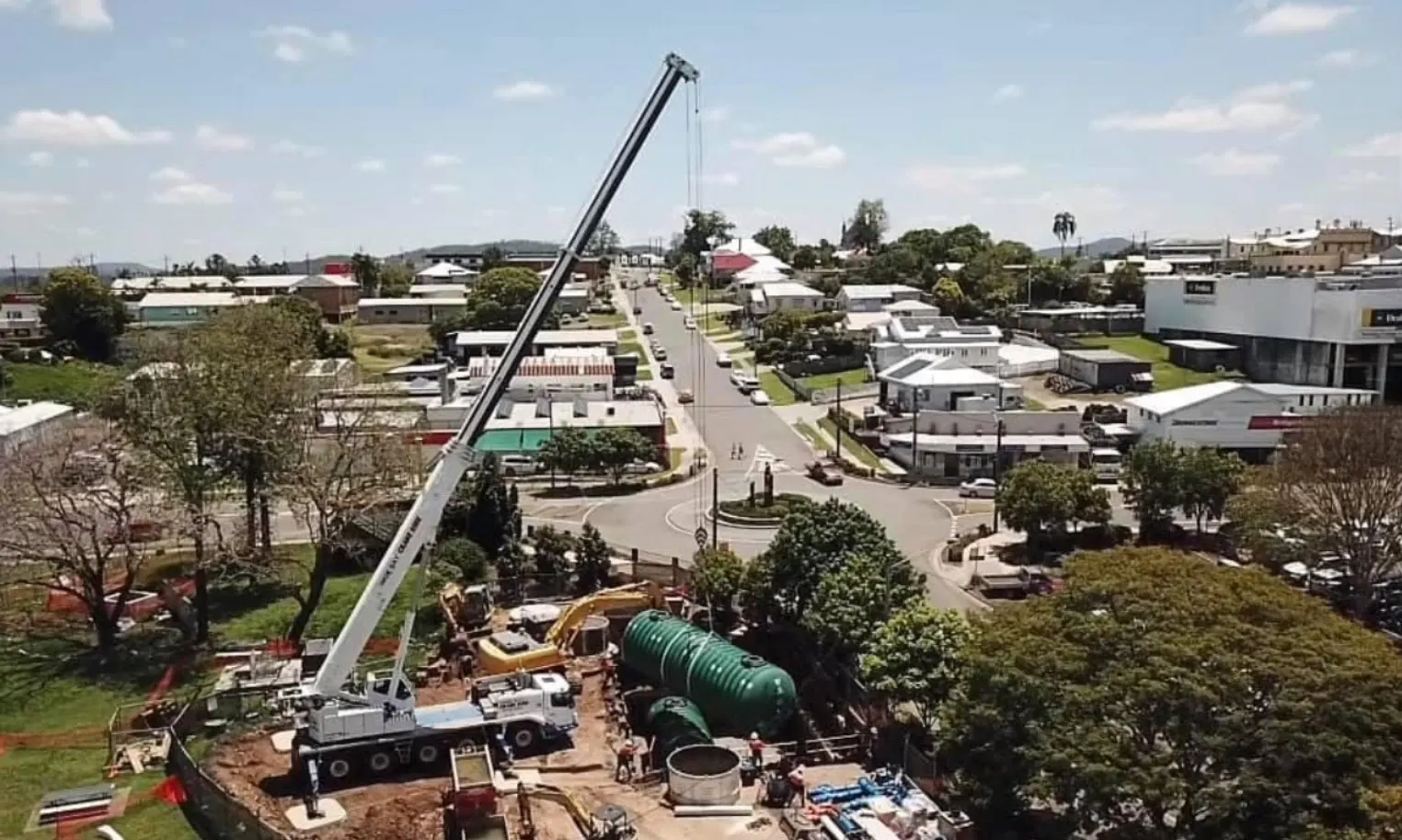 Aerial view of a mobile crane lowering green fibreglass water storage tanks into a shored excavation in central Gympie